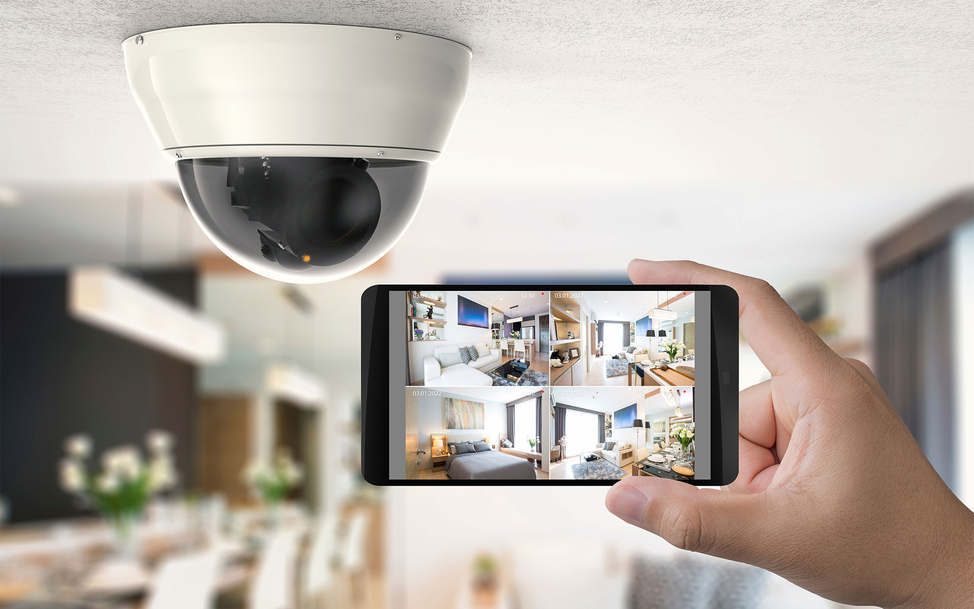 A close-up of an indoor security camera next to a person holding a mobile phone displaying video images of different rooms within a house