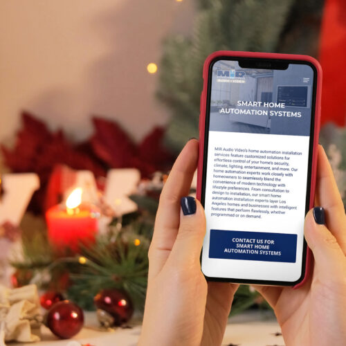 A woman in a festively decorated room holding a phone displaying MIR Audio Video's Smart Home Automation website page