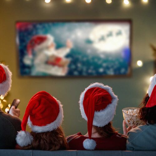 A view from the back of a family sitting on a couch, wearing Santa hats, eating popcorn, and watching a holiday movie in a festively decorated room