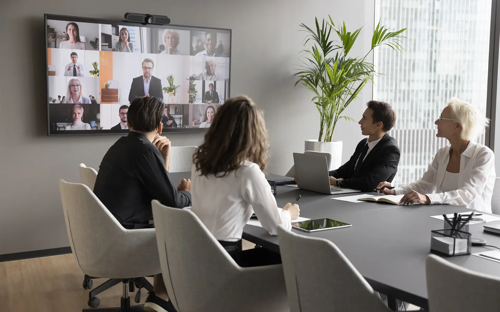 A view of a conference room interior featuring multiple employees conducting a video conference meeting on a wall-mounted screen with a web camera