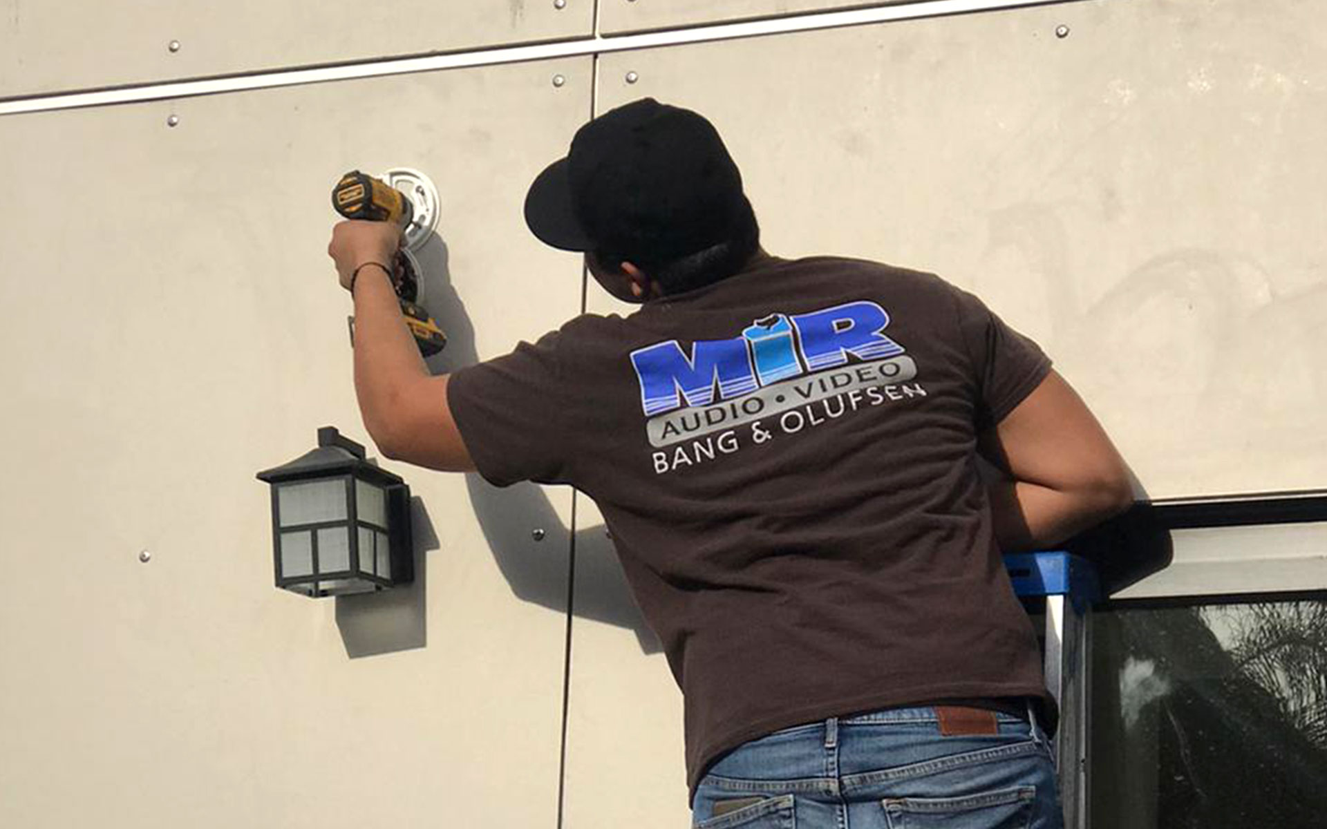 A MIR Audio Video smart home AV installer, Los Angeles, stands on a ladder, mounting a PTZ camera on the side of a home