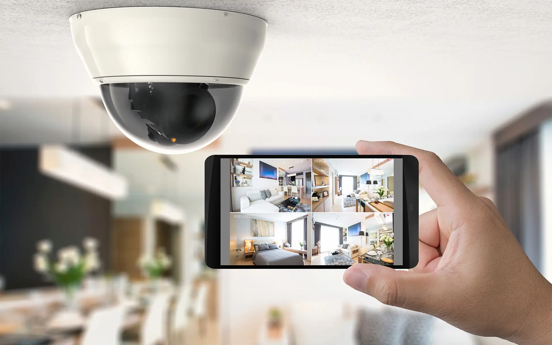 A hand holds a mobile phone under an indoor security camera with the screen displaying surveillance footage