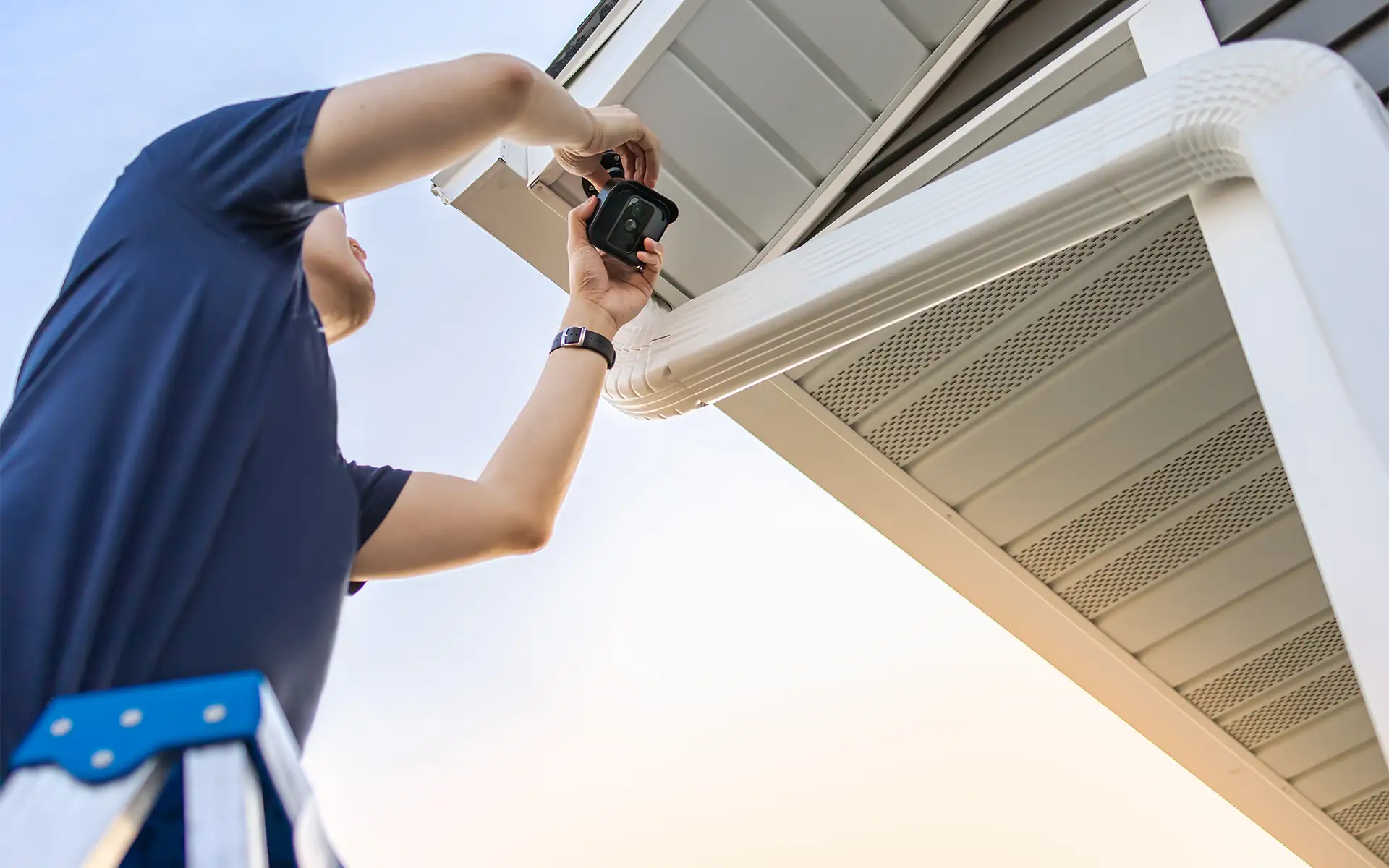 An audio video expert stands on a ladder while installing a home automation security camera on the exterior eave of a house