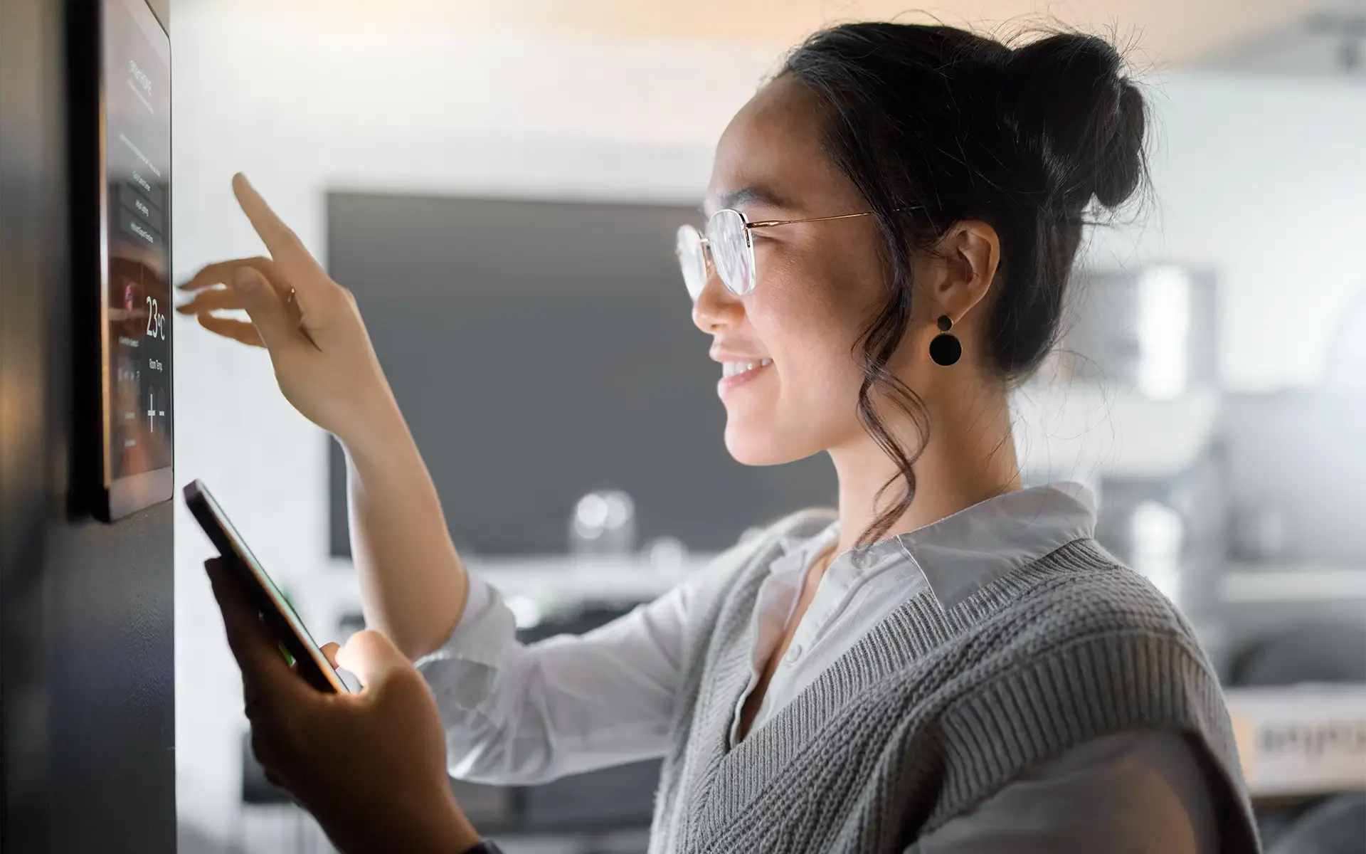 Side view of a woman holding a mobile phone while tapping the wall-mounted control panel of her home security and home automation system