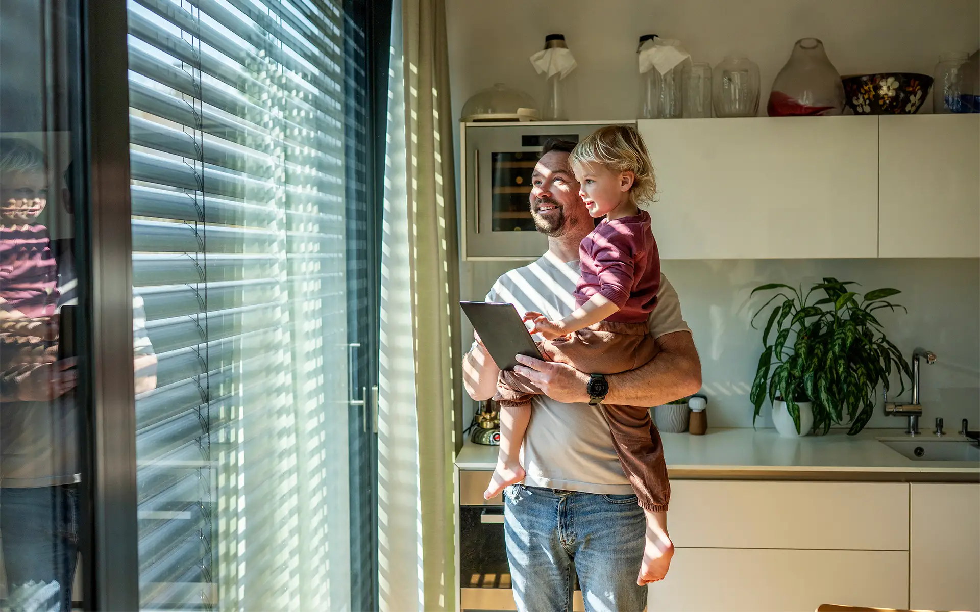 A father holds his toddler while standing next to a floor-to-ceiling window, demonstrating the shade control feature of their smart home automation control system