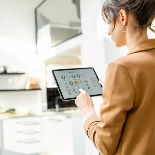 A woman stands in her sunny, white living room, using a tablet to control her smart home automation with the control system app