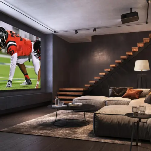 A basement home theater room with black walls and a tan sectional facing a very large projector screen showing a football game