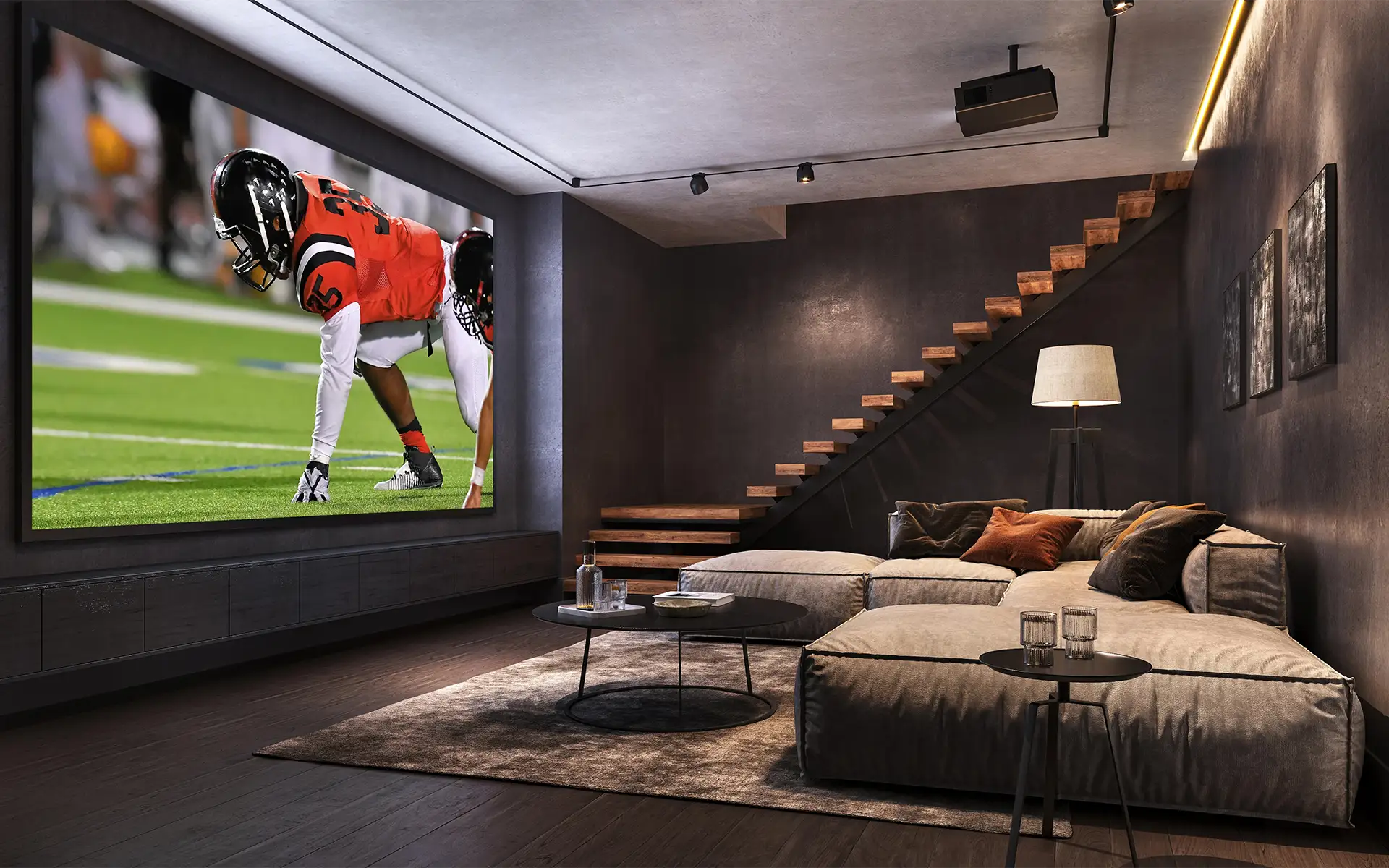 A basement home theater room with black walls and a tan sectional facing a very large projector screen showing a football game